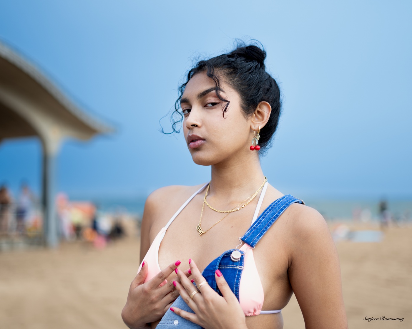 Woman with her hair in a bun and denim overalls looking toward the camera on a beach, a shade pavilion behind her.