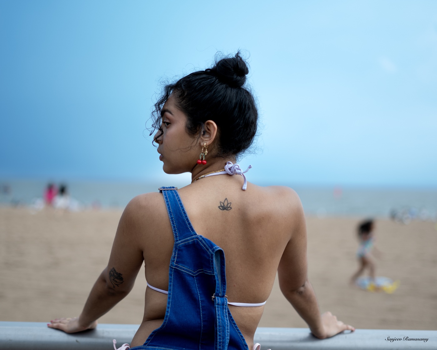 Woman with her hair in a bun, wearing denim overalls, seen from behind sitting on a railing above a sandy beach.