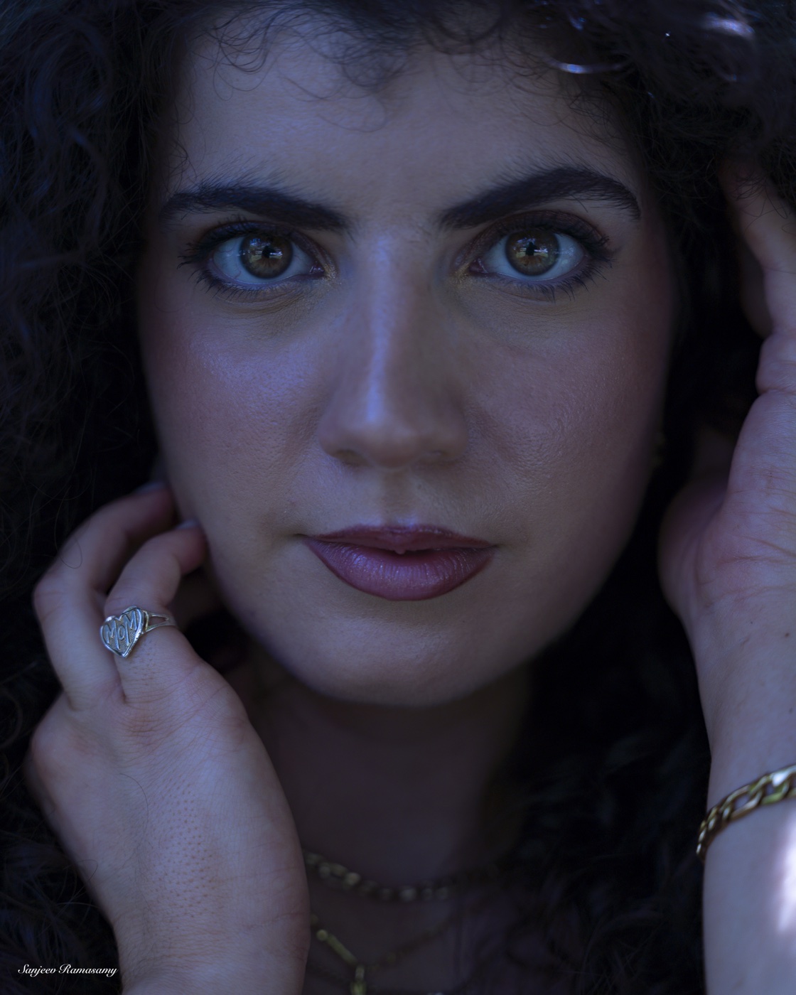 Close portrait of a woman with curly dark hair and light eyes, her hands framing her face against a dark background.
