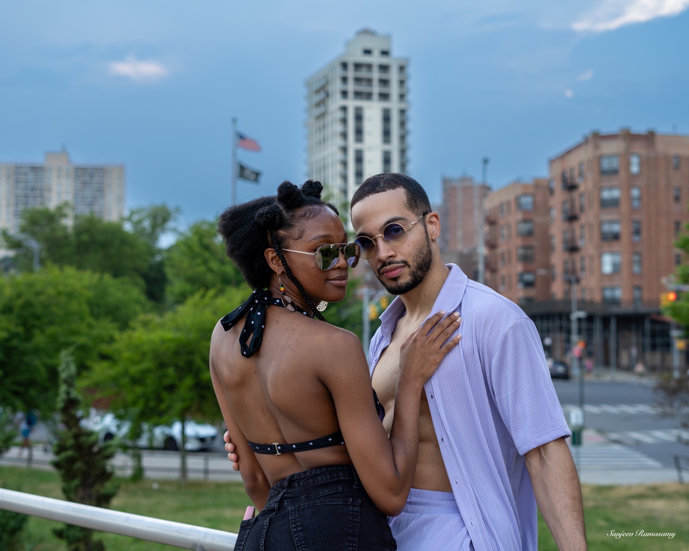 Man in a lavender shirt and a woman in a black halter top standing close by a railing, with apartment buildings and a flag behind them.