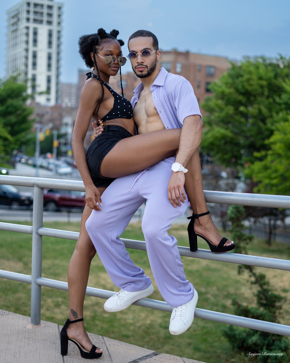 Man in a lavender outfit sitting on a park railing with a woman in black perched on his knee, high-rise buildings behind them.