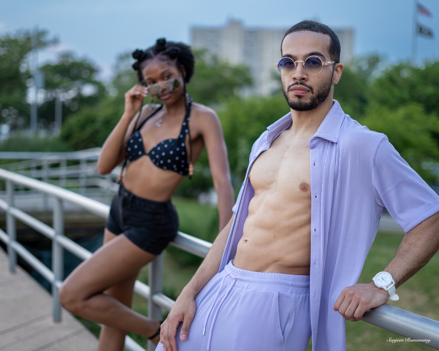 Man in an open lavender shirt and matching trousers and a woman in a polka-dot top and black shorts posing by a railing, both in sunglasses.