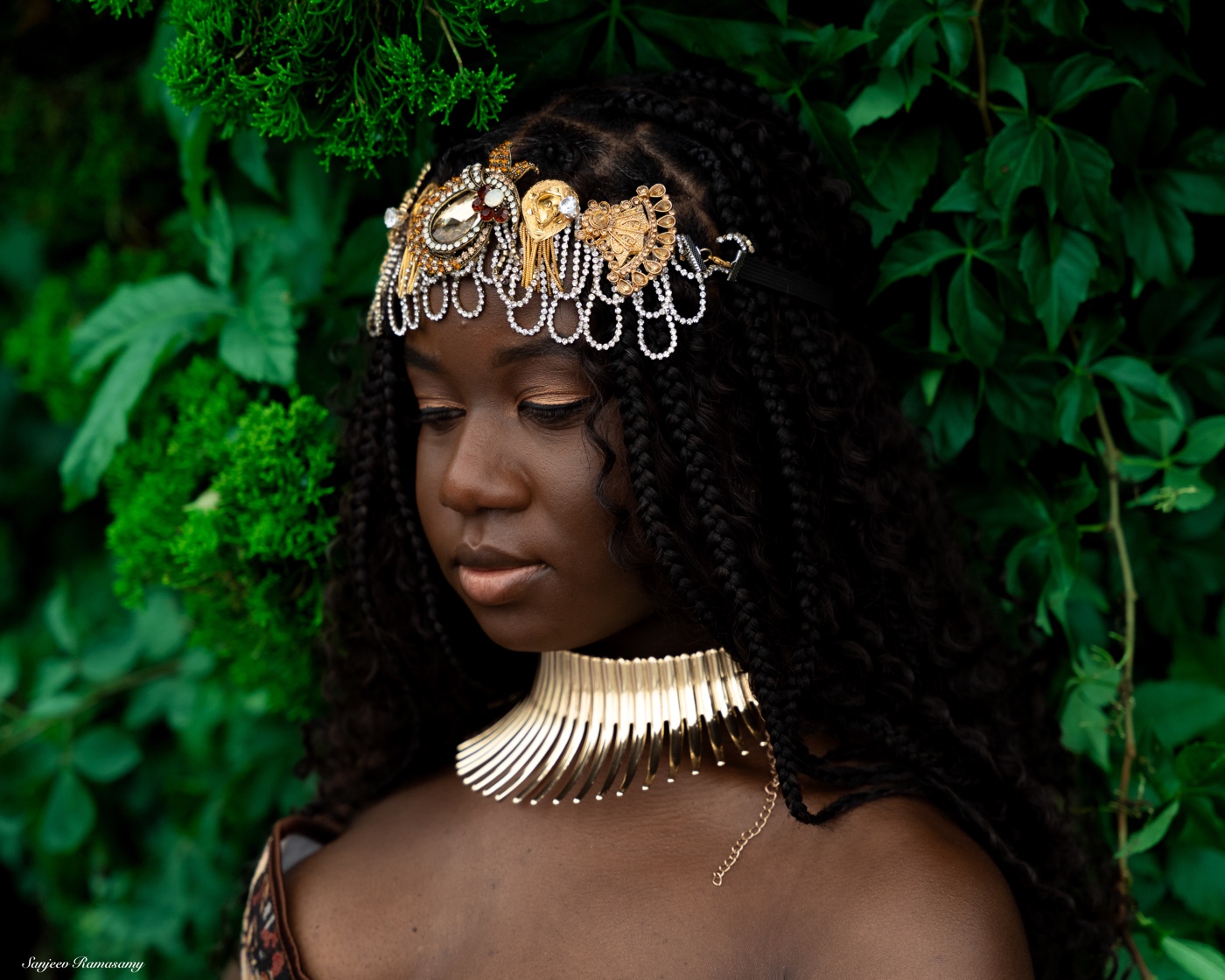 Woman in an ornate gold headpiece and collar necklace looking down, surrounded by dense green leaves.