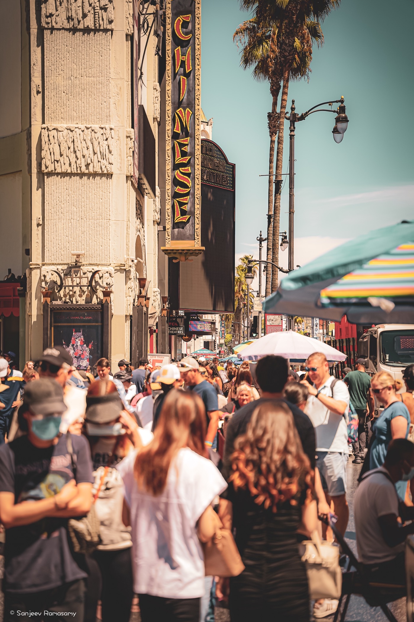 Crowd of people on a sunny street beneath palm trees and a tall vertical theatre sign.