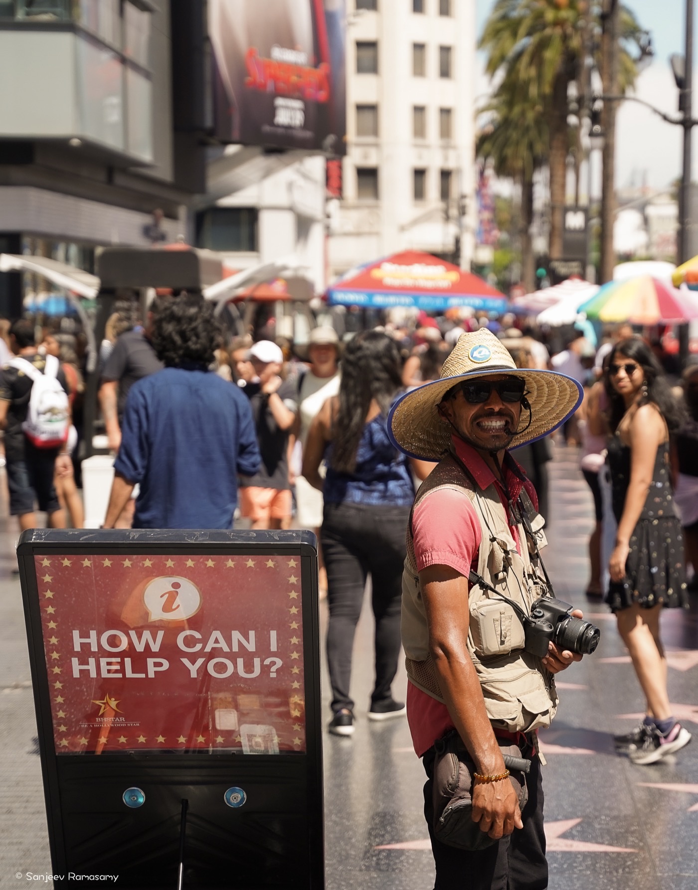 Smiling man in a wide-brimmed hat with a camera on a crowded sidewalk, beside a red kiosk reading 'How can I help you?'