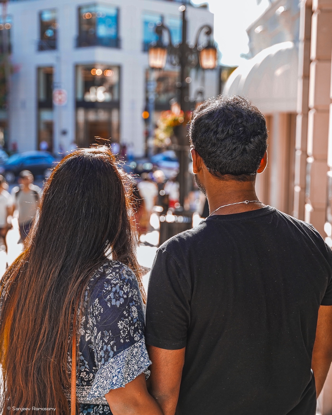 A couple seen from behind, a woman with long hair and a man in a black T-shirt, standing close on a busy city street.