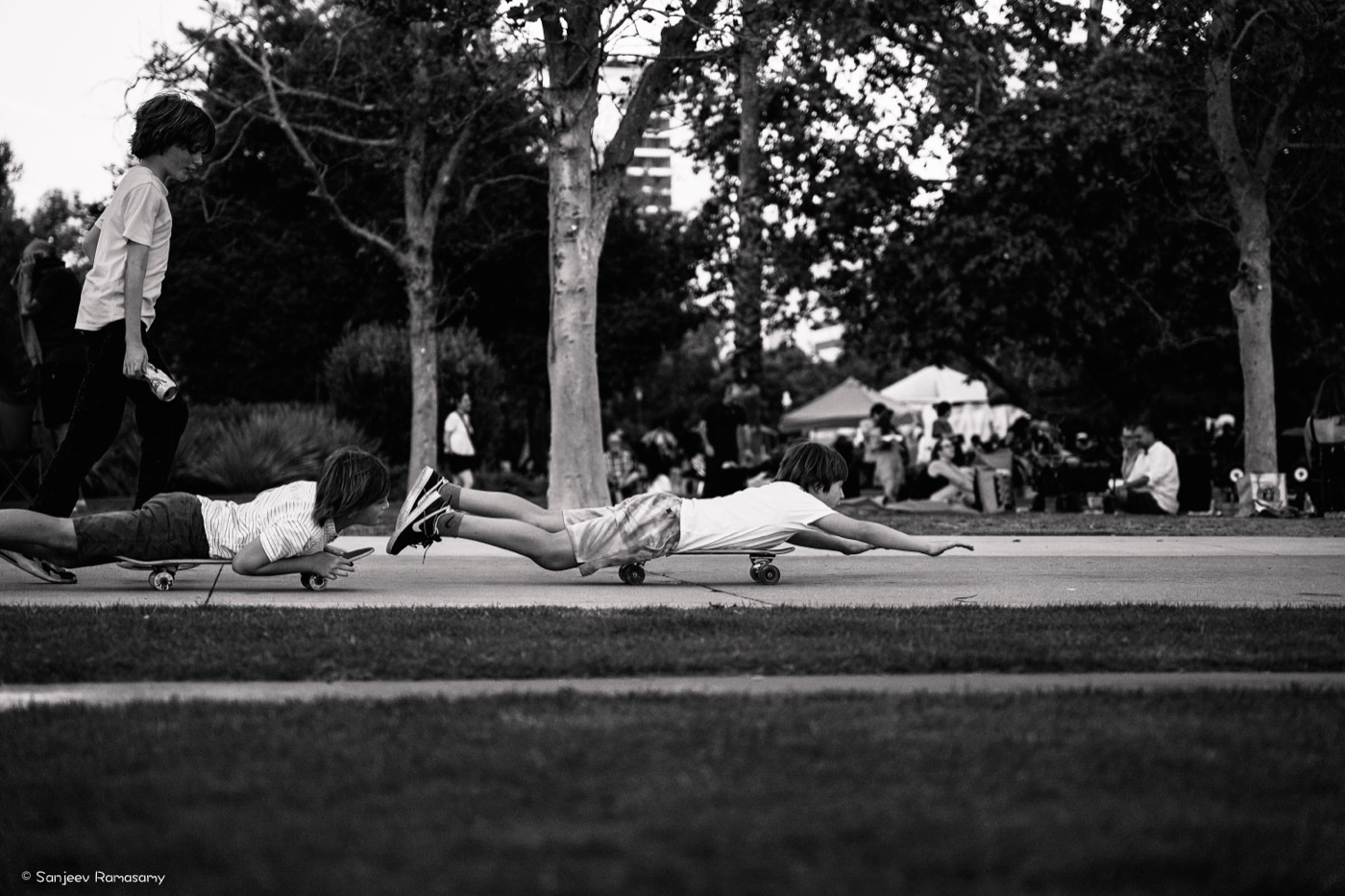 Black-and-white photo of people stretched out on skateboards across a park path under large trees.