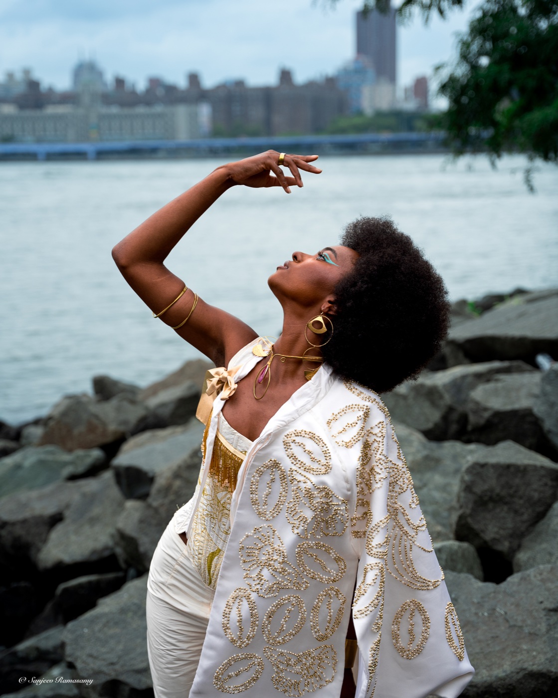 Woman in a white-and-gold jacket posing on shoreline rocks with one arm raised, a city skyline across the water behind her.