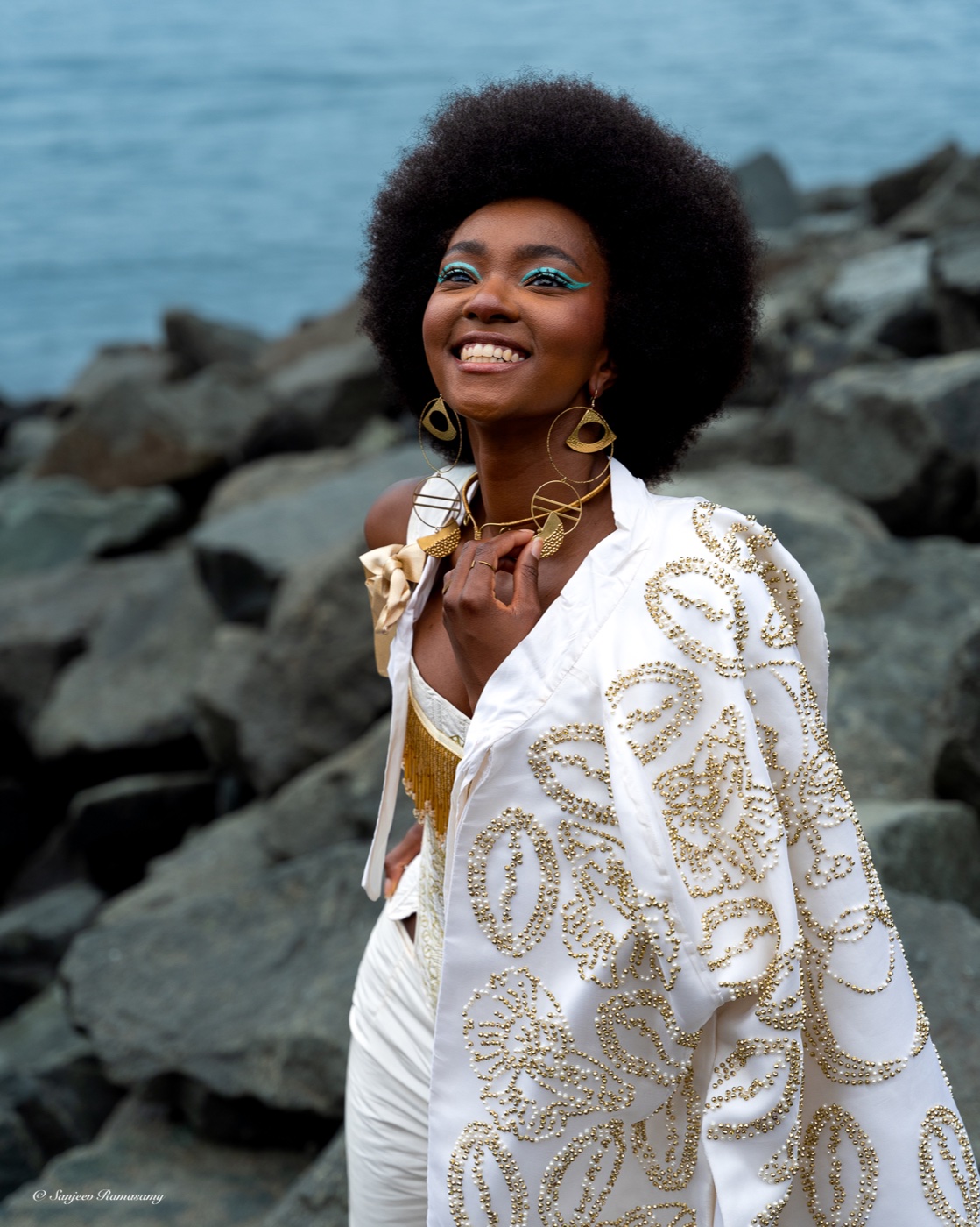 Smiling woman with voluminous natural hair in a white-and-gold jacket, standing on shoreline rocks and looking upward.