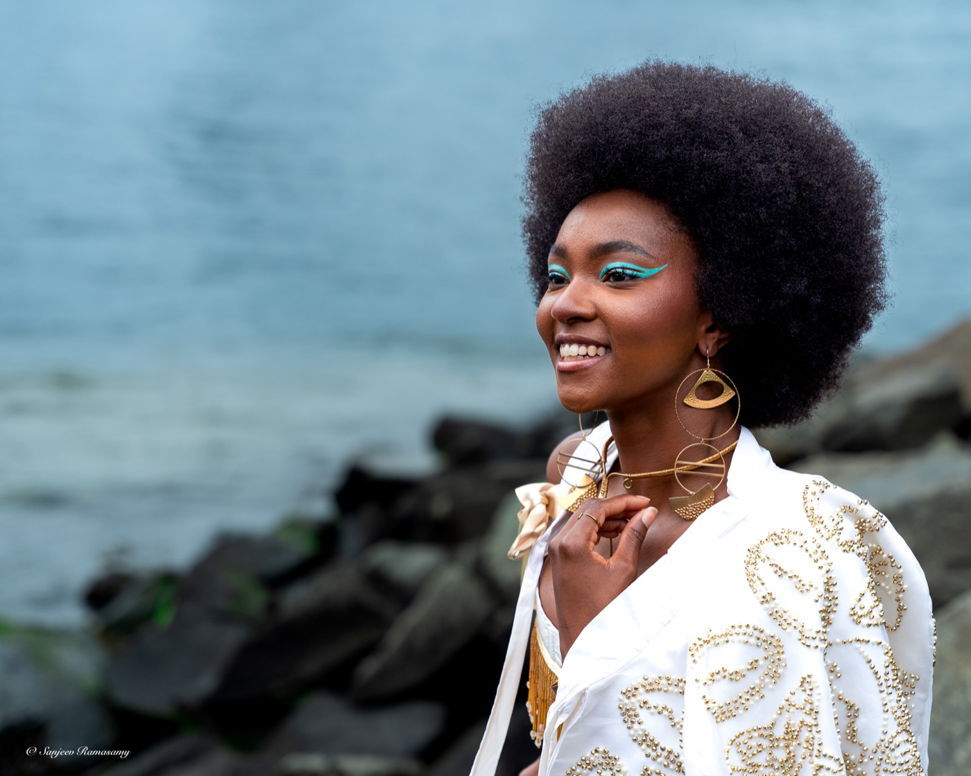 Laughing woman with voluminous natural hair, turquoise eye makeup and gold earrings, in a white-and-gold jacket by the water.