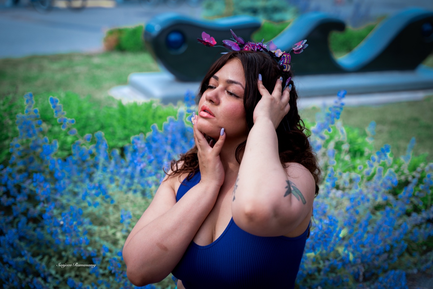 Woman with purple flowers in her hair posing among blue blossoms in a garden, hands raised to her face.