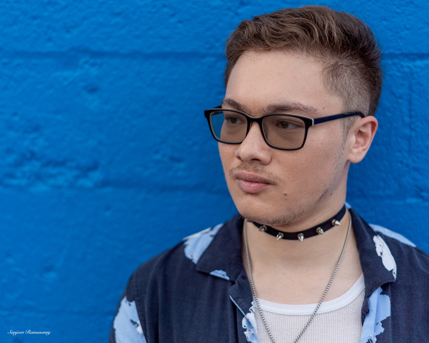 Portrait of a young man with glasses, a studded choker and a patterned shirt against a bright blue wall.