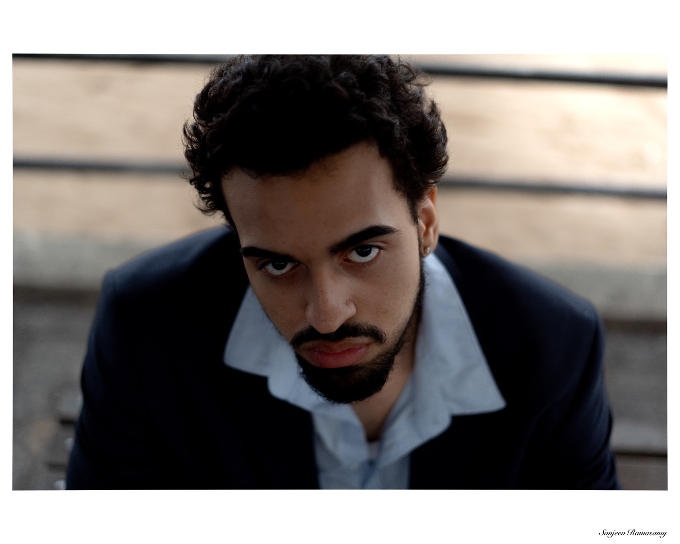 Close portrait of a man with curly hair and a beard in a dark jacket and open-collared white shirt, gazing into the camera.
