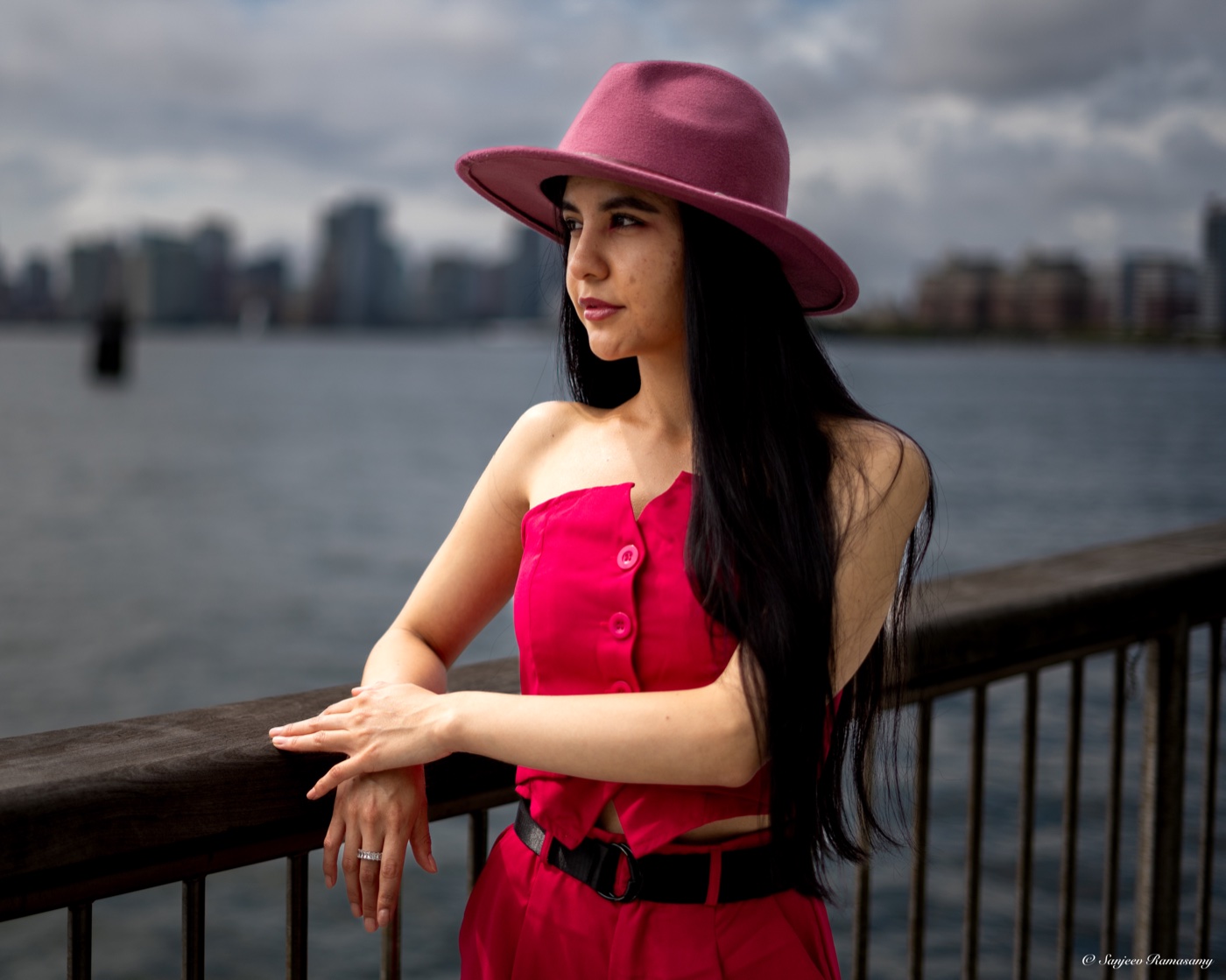 Woman in a pink hat and red outfit leaning on a waterfront railing, looking away toward the skyline.