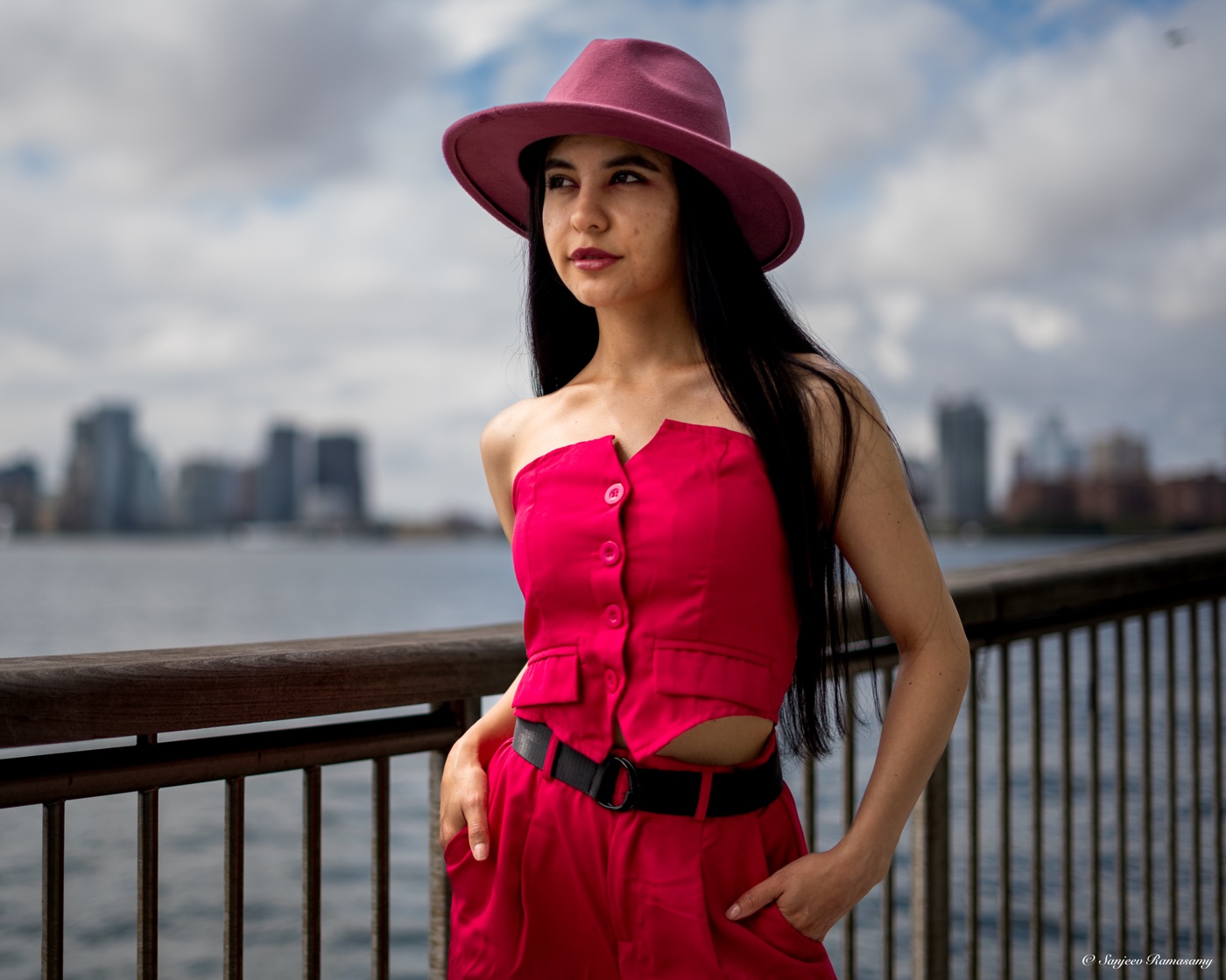 Woman in a wide-brimmed pink hat and a red strapless top standing by a waterfront railing, with a city skyline across the water.