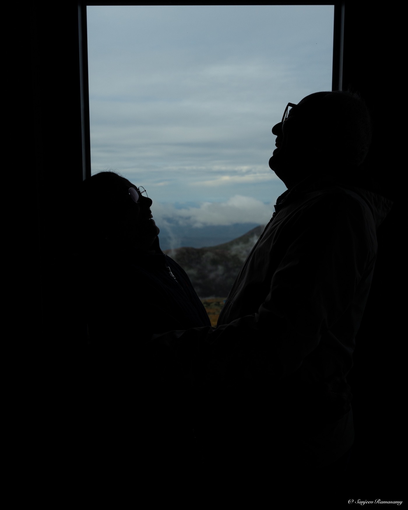 Two people in near-silhouette beside a window that looks out over hazy mountains.