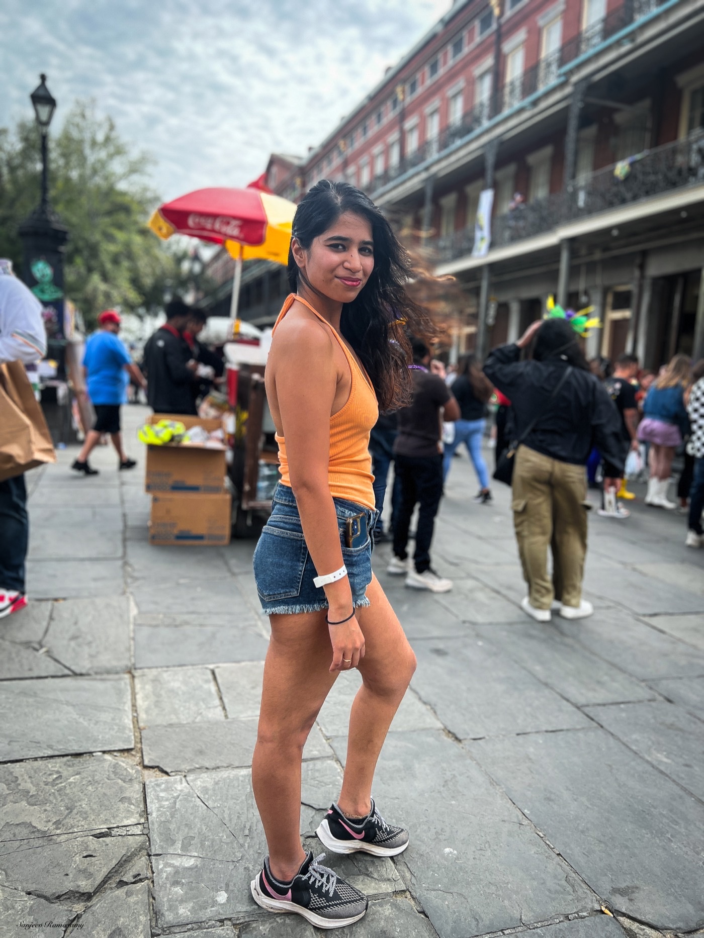 Woman in an orange top and denim shorts walking along a busy pedestrian street lined with balconied buildings and vendors.