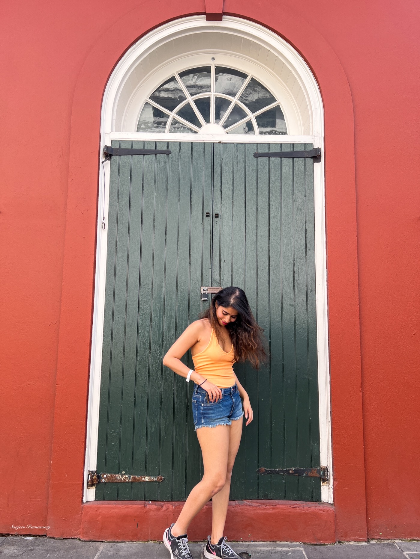 Woman in an orange top and denim shorts posing in front of a tall arched green door set in a red wall.