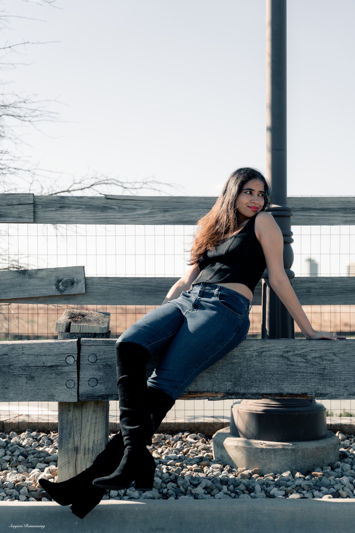 Woman in a black top, jeans and knee-high boots leaning back on a weathered wooden fence, looking over her shoulder.