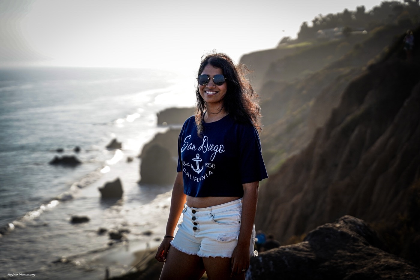 Smiling woman in sunglasses and a navy San Diego T-shirt standing on a cliff above the ocean, backlit by the late sun.