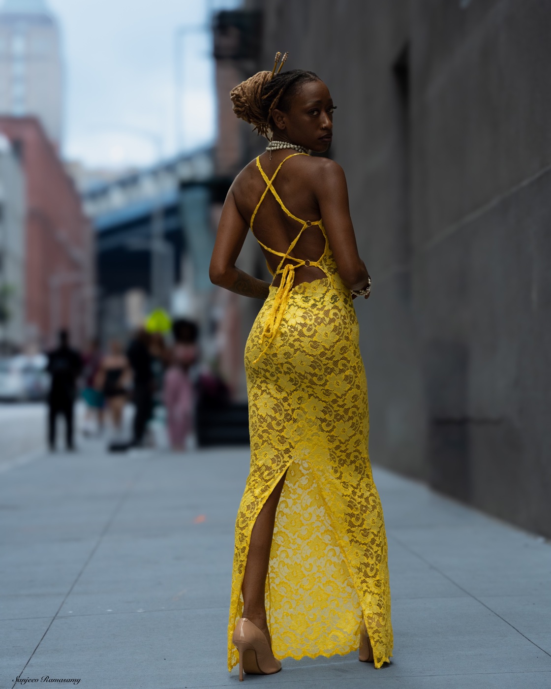 Woman in a yellow lace gown looking back over her shoulder on a city sidewalk, with a bridge and buildings behind her.
