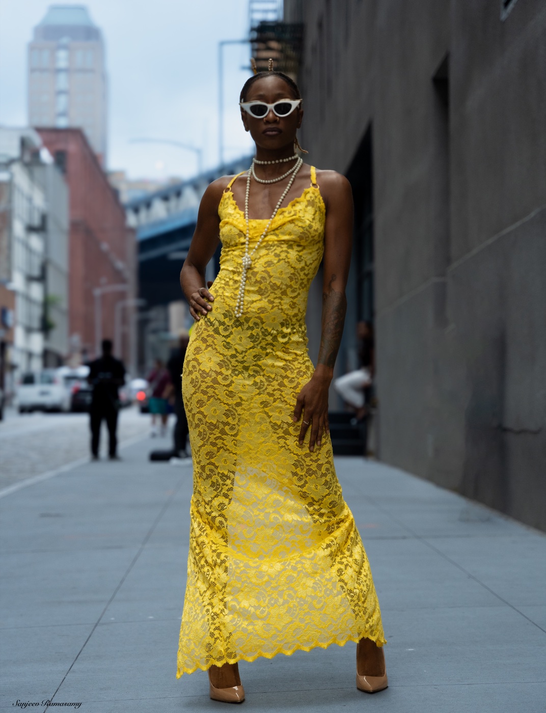 Woman in white sunglasses, layered necklaces and a floor-length yellow lace gown standing on a city sidewalk.