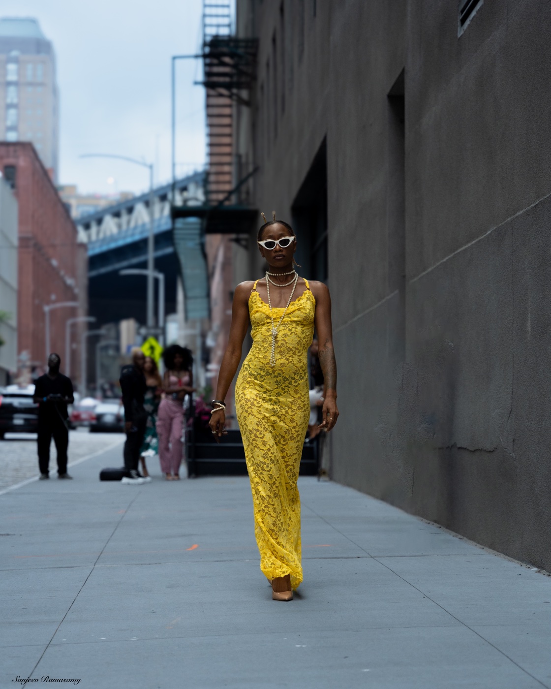 Woman in white sunglasses and a yellow lace gown walking toward the camera along a city sidewalk.