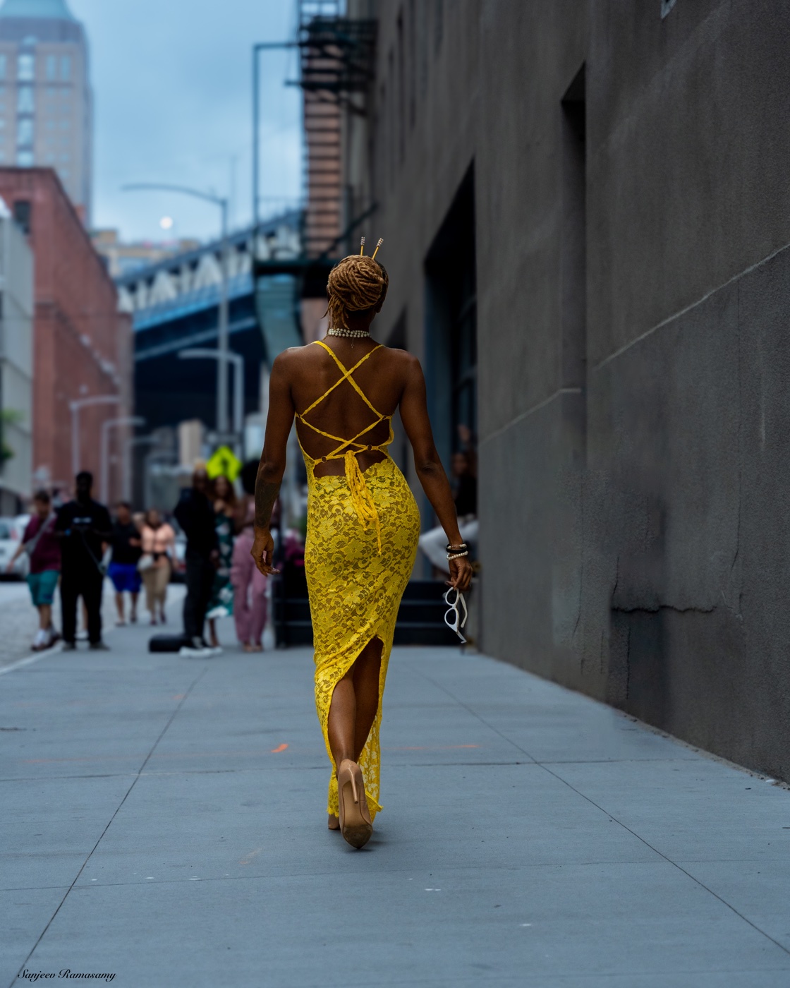 Woman in a yellow lace gown with a crossed-strap back walking away down a city sidewalk between tall buildings.